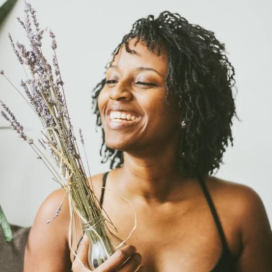 Smiling Black woman with sisterlocks holding a small glass vase of dried lavender.