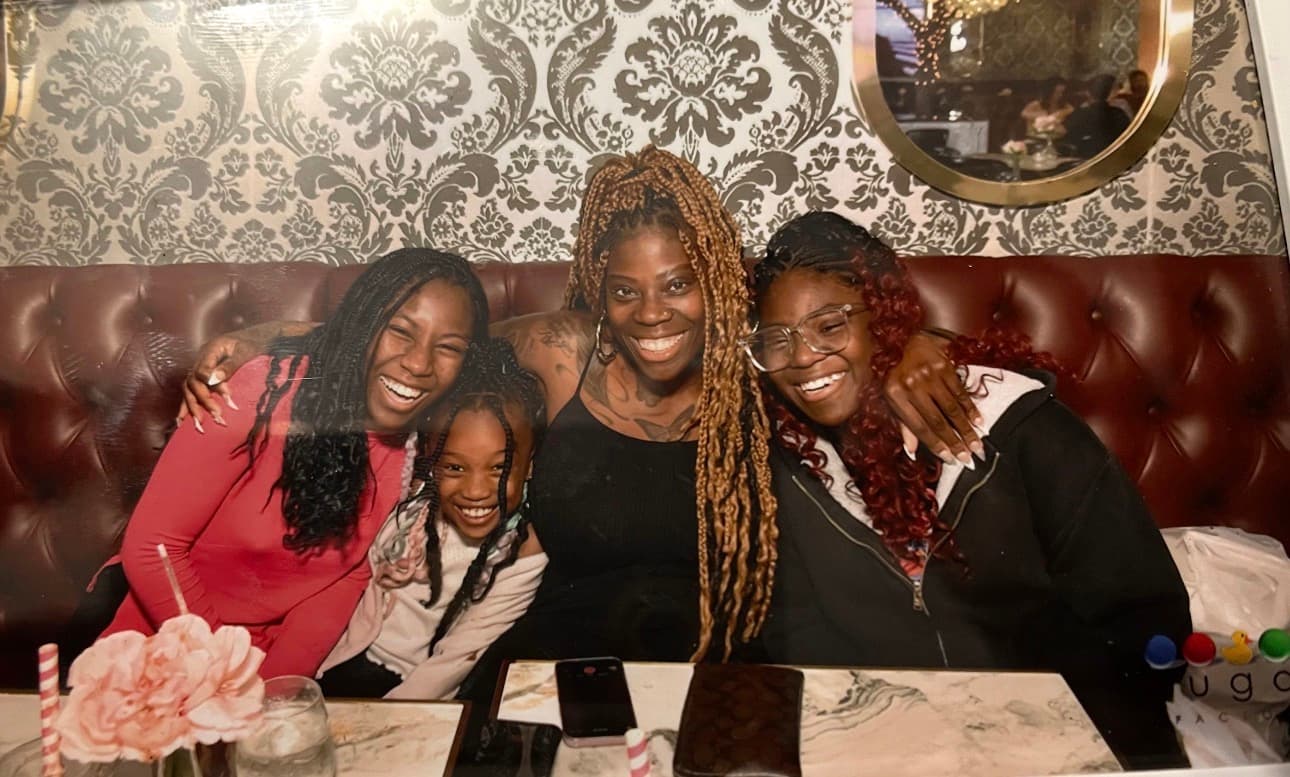 Four smiling Black females pose together on a tufted leather booth against patterned wallpaper.